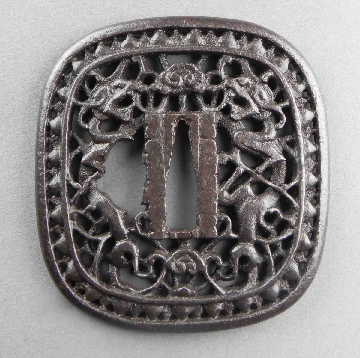 Tsuba (garde de sabre japonais) en métal de forme carrée avec des motifs ajourés complexes.