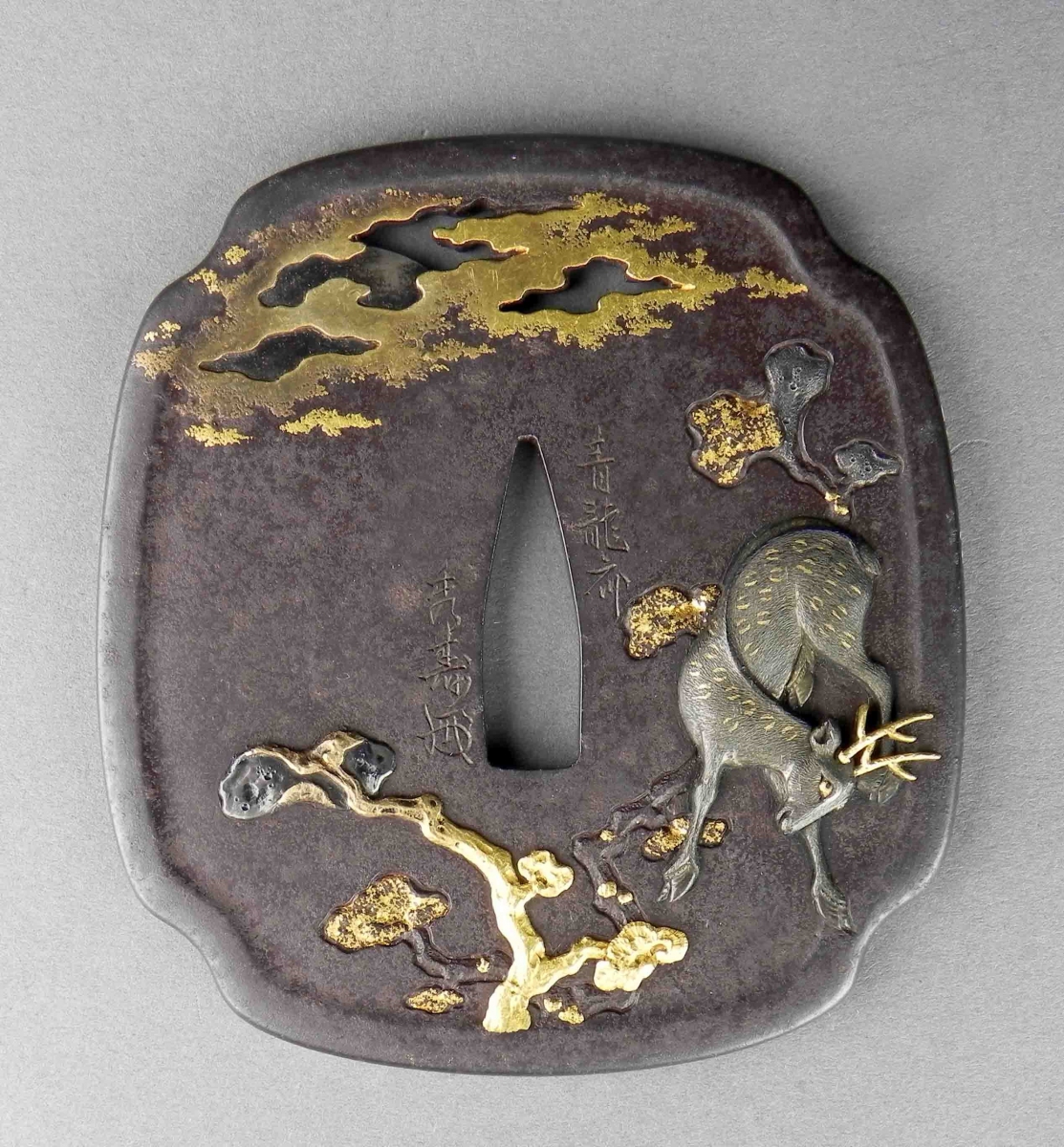 Tsuba en fer avec des accents dorés représentant un cerf sous un arbre et des nuages sur un fond de patine sombre.