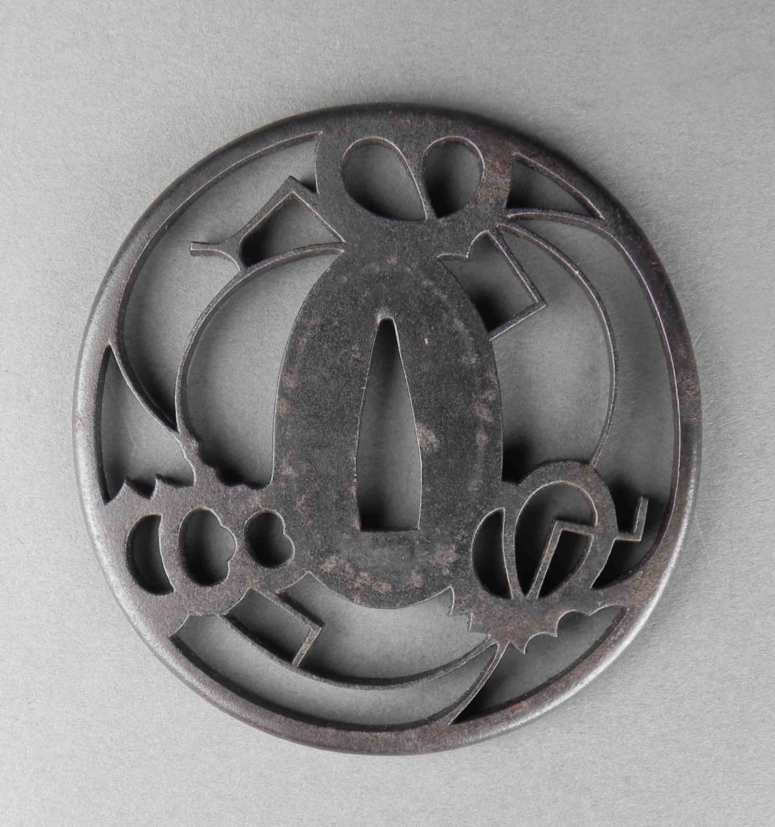 Tsuba ronde en métal foncé avec des formes abstraites découpées autour de l'ouverture ovale centrale.