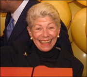 An elderly woman with short gray hair smiling, wearing a black top and pearl earrings.