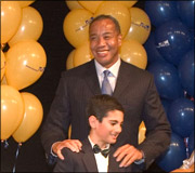 A man in a dark suit and tie stands behind a boy in a tuxedo with a bow tie, with yellow and blue balloons in the background.