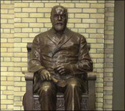 Bronze statue of a seated man with a beard wearing a suit, against a yellow brick wall.