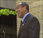 Man in a dark suit and tie smiling, standing in front of a yellow brick wall.