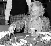 An elderly woman sitting at a table, smiling and cutting a cake with a knife.