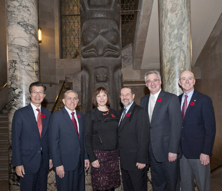 Six formally dressed people standing indoors in front of a tall totem pole and marble columns.