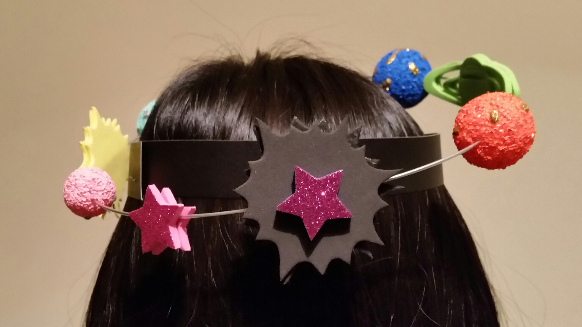 A headband featuring glittery cosmic shapes and small planets on springs above a person's dark hair.
