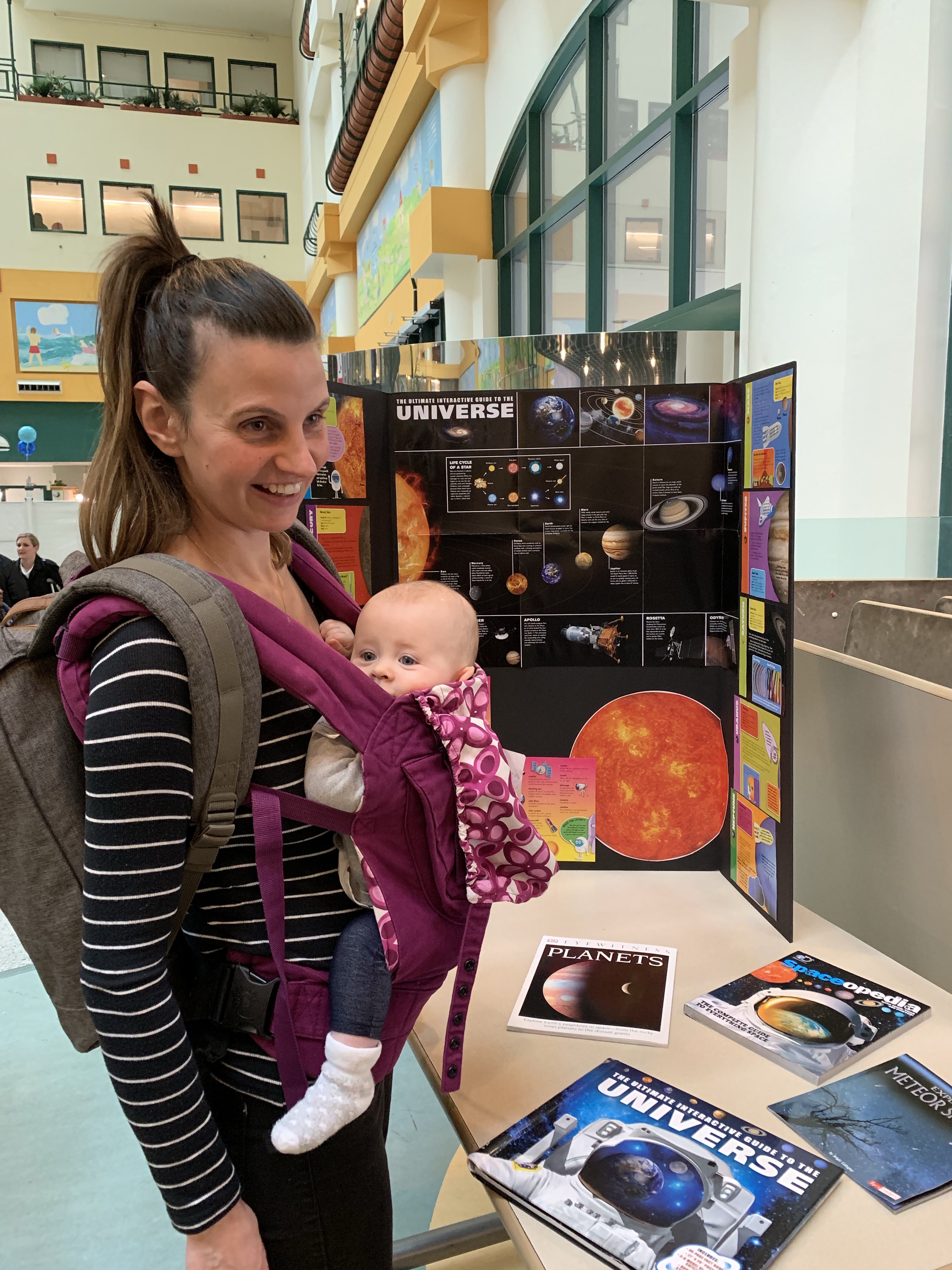 A woman carrying a baby in a front carrier stands next to a display of space-themed photos and illustrations.