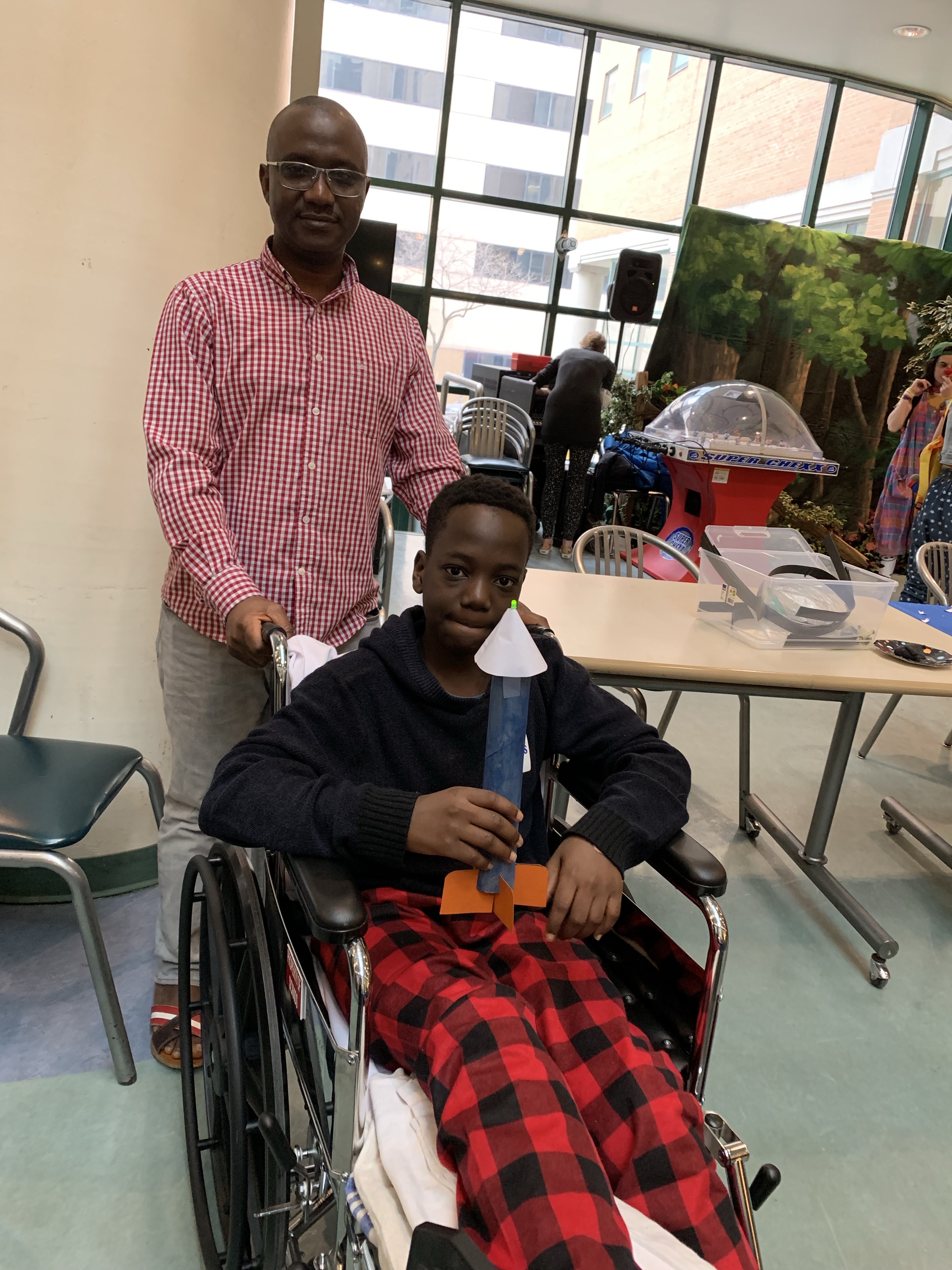 A man in a pink shirt stands behind a smiling young person in a wheelchair holding a model rocket.