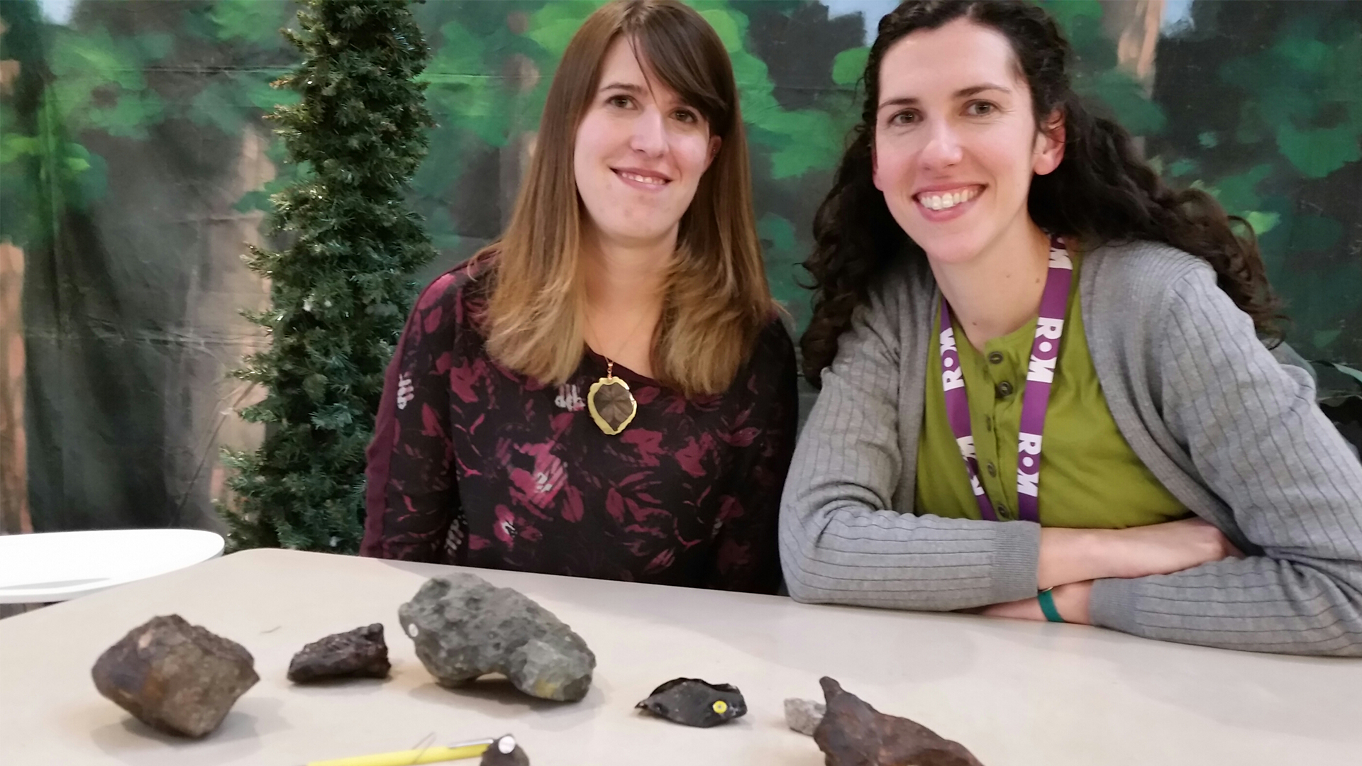 Two women sitting behind a table with five various fossil specimens displayed on it.