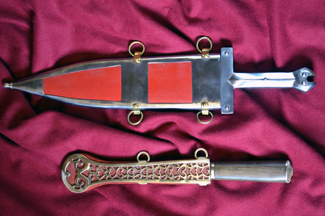 A dagger with a decorated sheath featuring red rectangles and gold rings next to an ornately carved brass sheath.