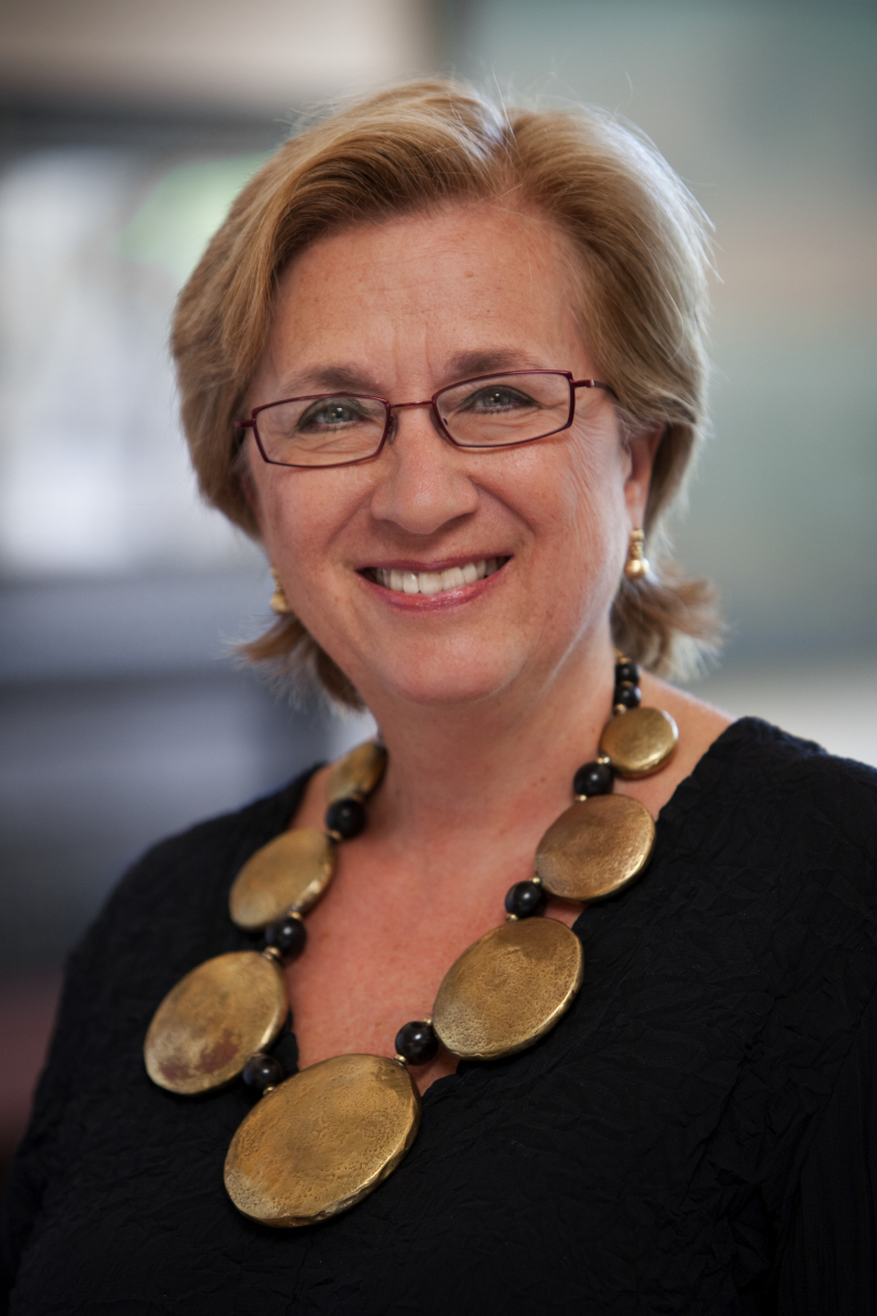 Portrait of a smiling woman with short blonde hair, glasses, and a large circular necklace on a black top.