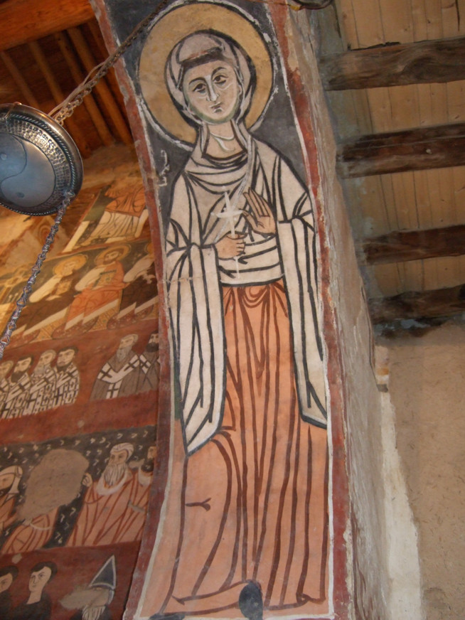 A painted religious figure with a halo, wearing a long orange robe and white cloak, holding a scroll, on an interior wall.