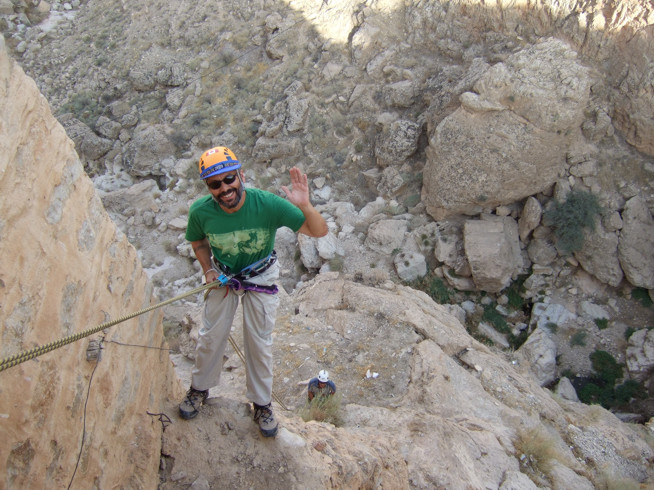 Man wearing a green shirt, helmet, and climbing harness giving a thumbs-up while rappelling down a rocky cliff.