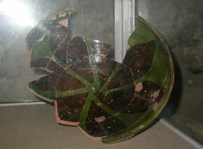A broken green ceramic bowl with dark brown spots displayed behind glass.