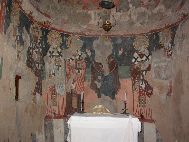 Faded religious mural depicting four haloed figures in robes inside a curved, aged wall space with an altar table covered by a white cloth in front.