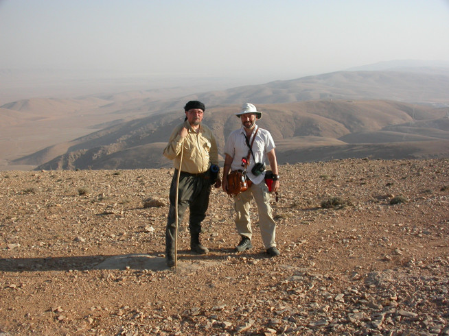 Two men standing on a rocky hilltop with a wide, arid landscape of rolling hills in the background.
