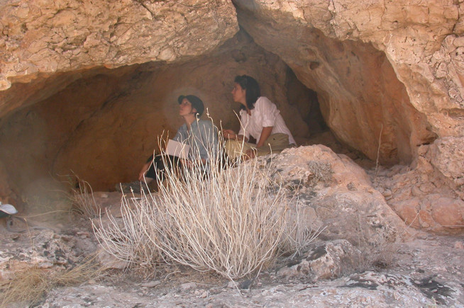 Two people sitting inside a rocky cave partially hidden by dry shrubbery in the foreground.