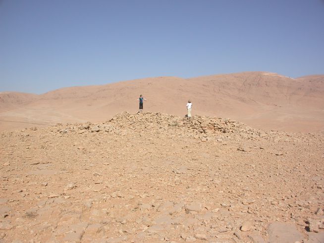 Two people standing on a small rocky mound in a barren desert landscape with hills in the background.