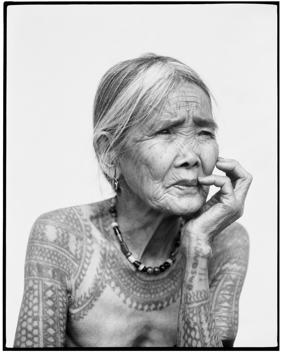 Elderly woman with traditional tattoos on her arms and upper chest, resting her chin on her hand.