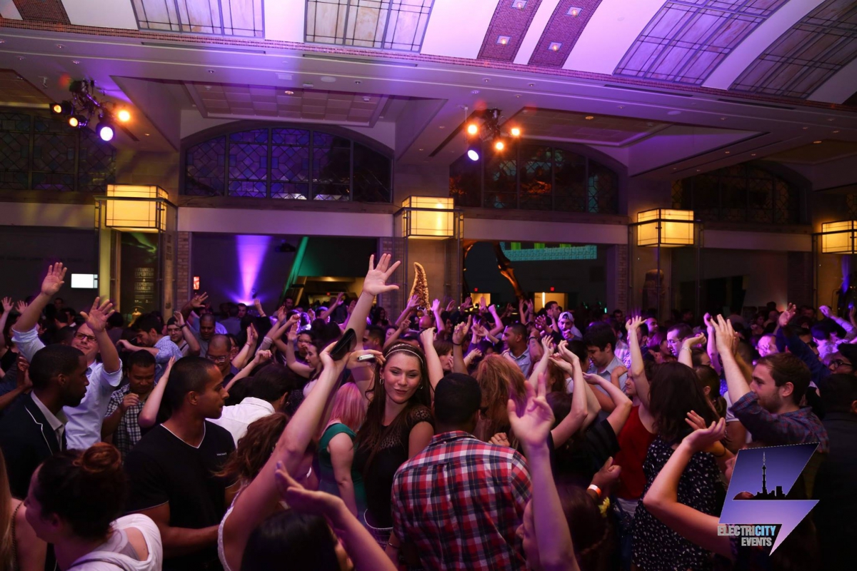 Crowd of people dancing and raising their hands in a large indoor venue with arched ceilings and purple lighting.