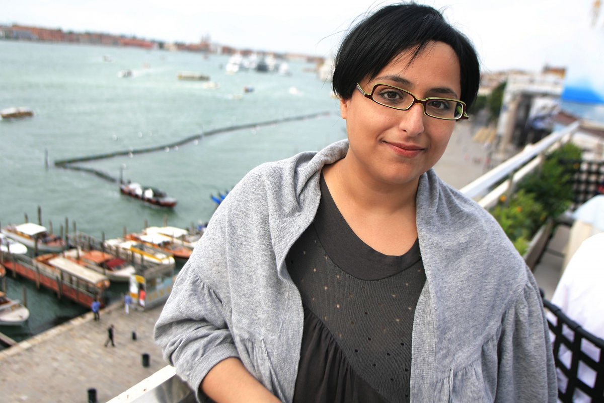 Woman with glasses wearing a gray shawl standing near a waterfront with boats and buildings in the background.
