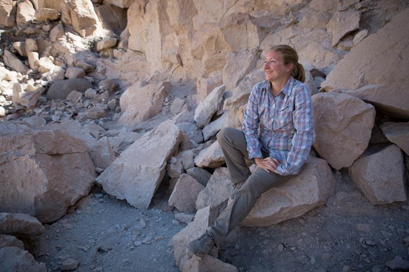 A person wearing a plaid shirt and gray pants sits on large rocks in a rocky, dry outdoor setting.