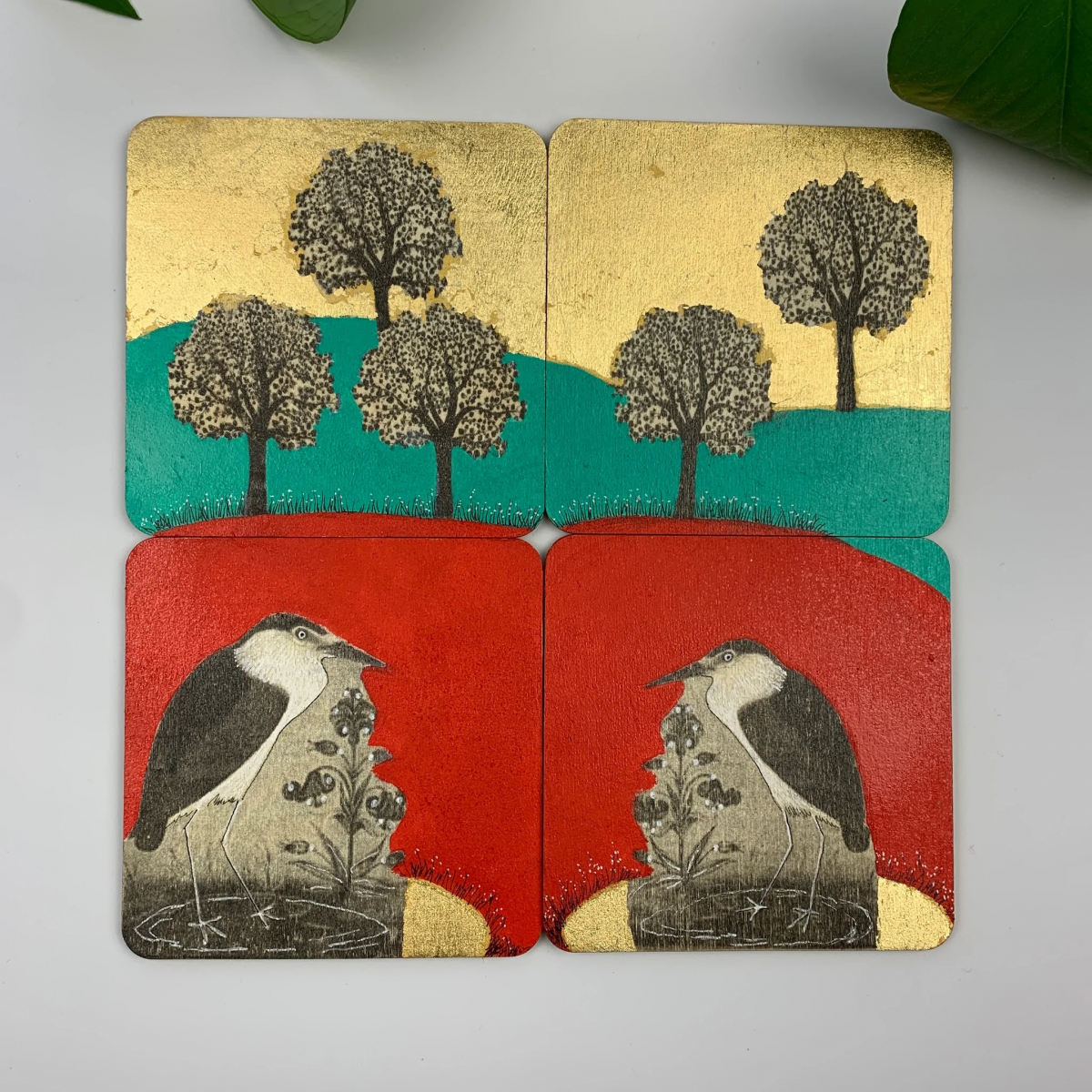Four square coasters with textured art: the top two show black trees on yellow and green backgrounds; the bottom two show black and white birds on red and yellow backgrounds.