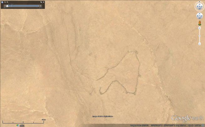 Google Earth satellite map showing an irregularly shaped stone enclosure in a sandy area with faint tracks nearby.