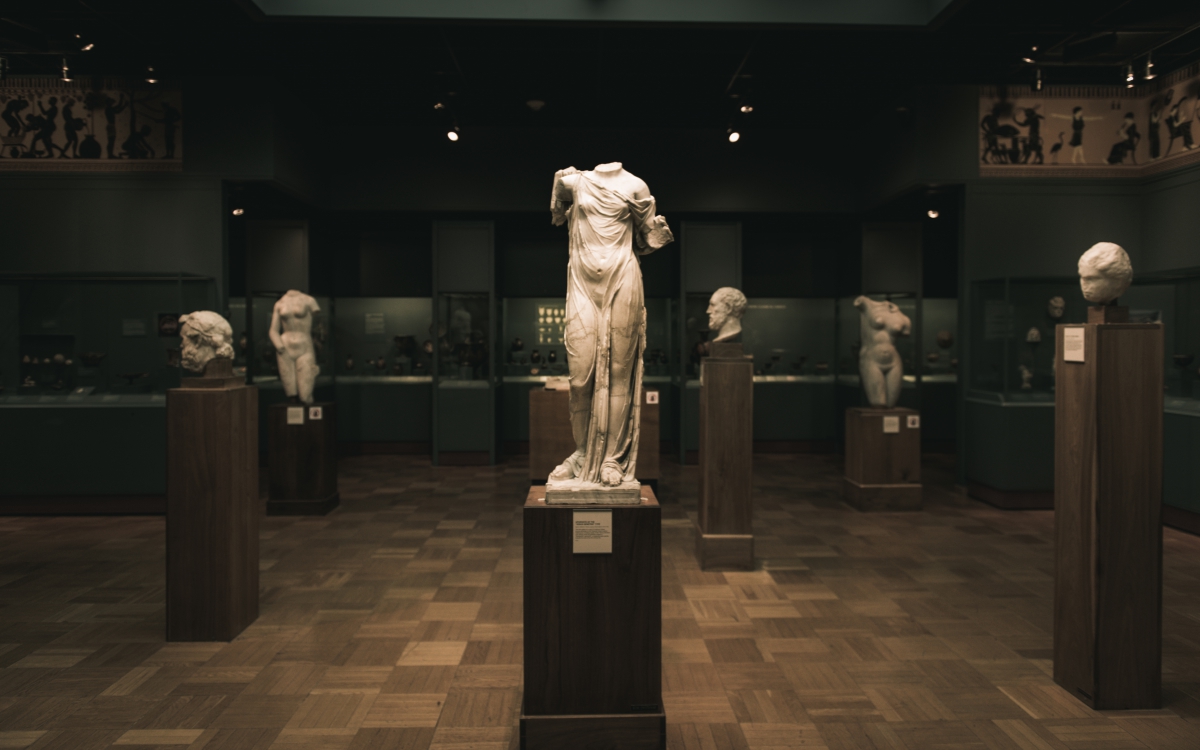 A museum gallery with a central statue of a headless, armless classical torso on a pedestal, surrounded by busts on pedestals.