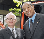 Portrait of two men smiling, one wearing glasses and a grey suit, the other in a dark suit with a pin on his lapel.