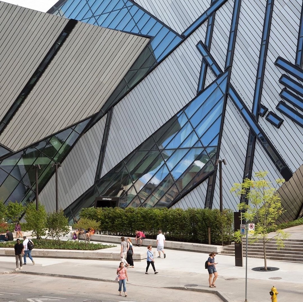 Modern angular building facade with large reflective glass panels and people walking on the sidewalk in front.