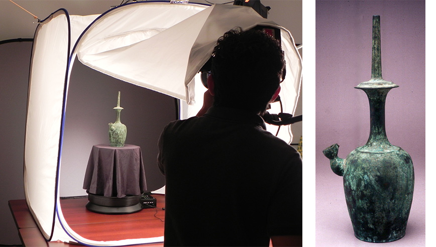 Photograph showing a person silhouetted against a bright light photographing a greenish bronze artifact shaped like a vessel with a tall neck and cross-shaped top.