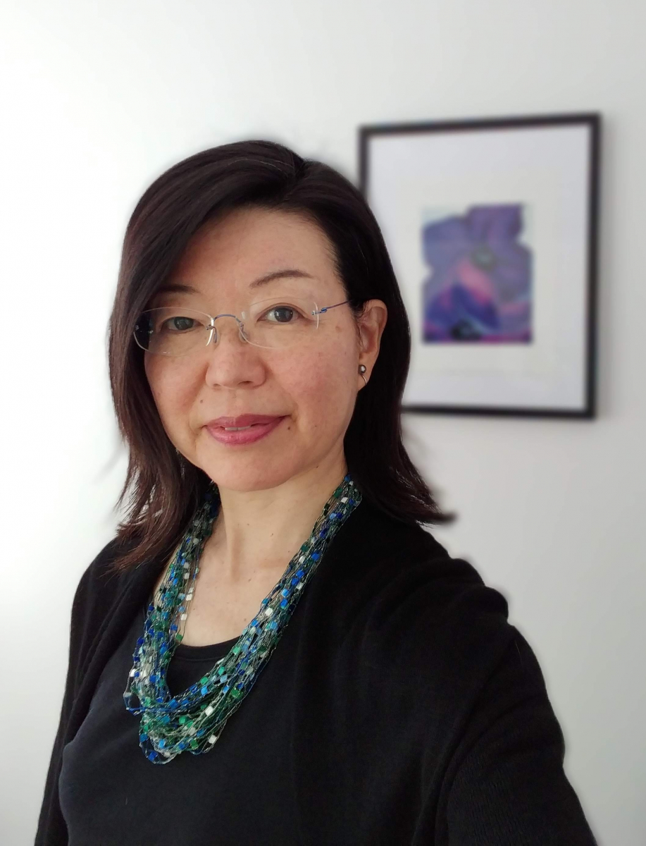 Portrait of an Asian woman with glasses wearing a black top and a layered blue-green beaded necklace, standing in front of a framed abstract artwork.