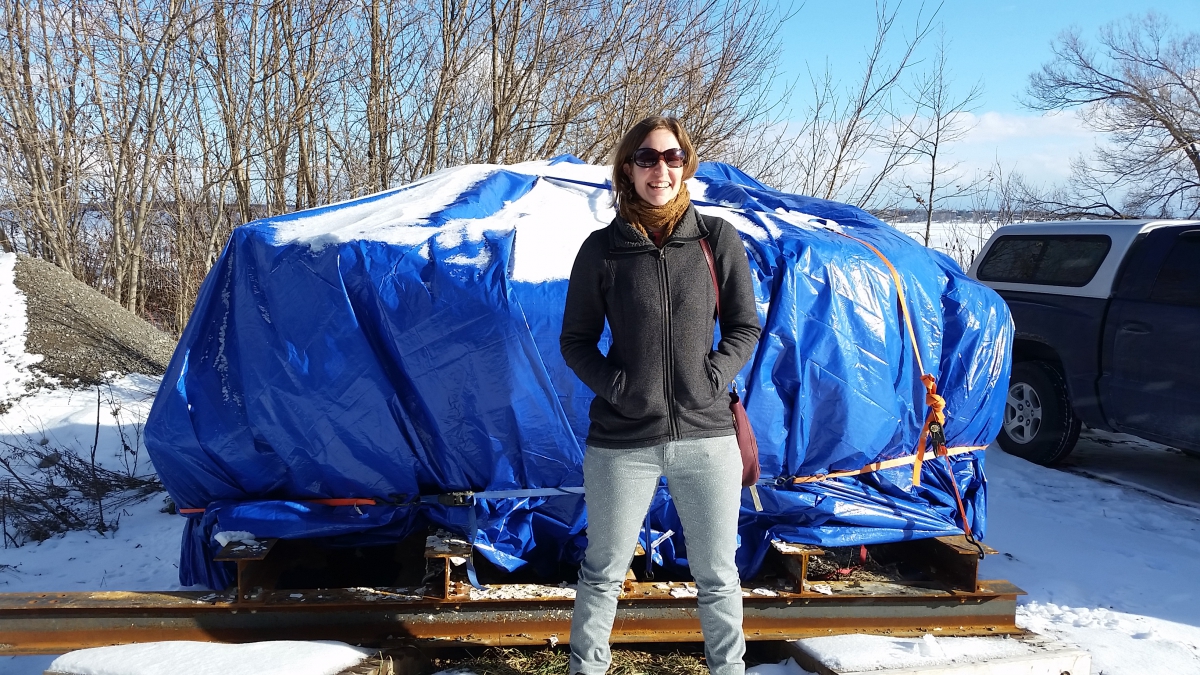 Man wearing sunglasses and a gray jacket standing in front of a large object covered by a blue tarp outdoors in a snowy setting.