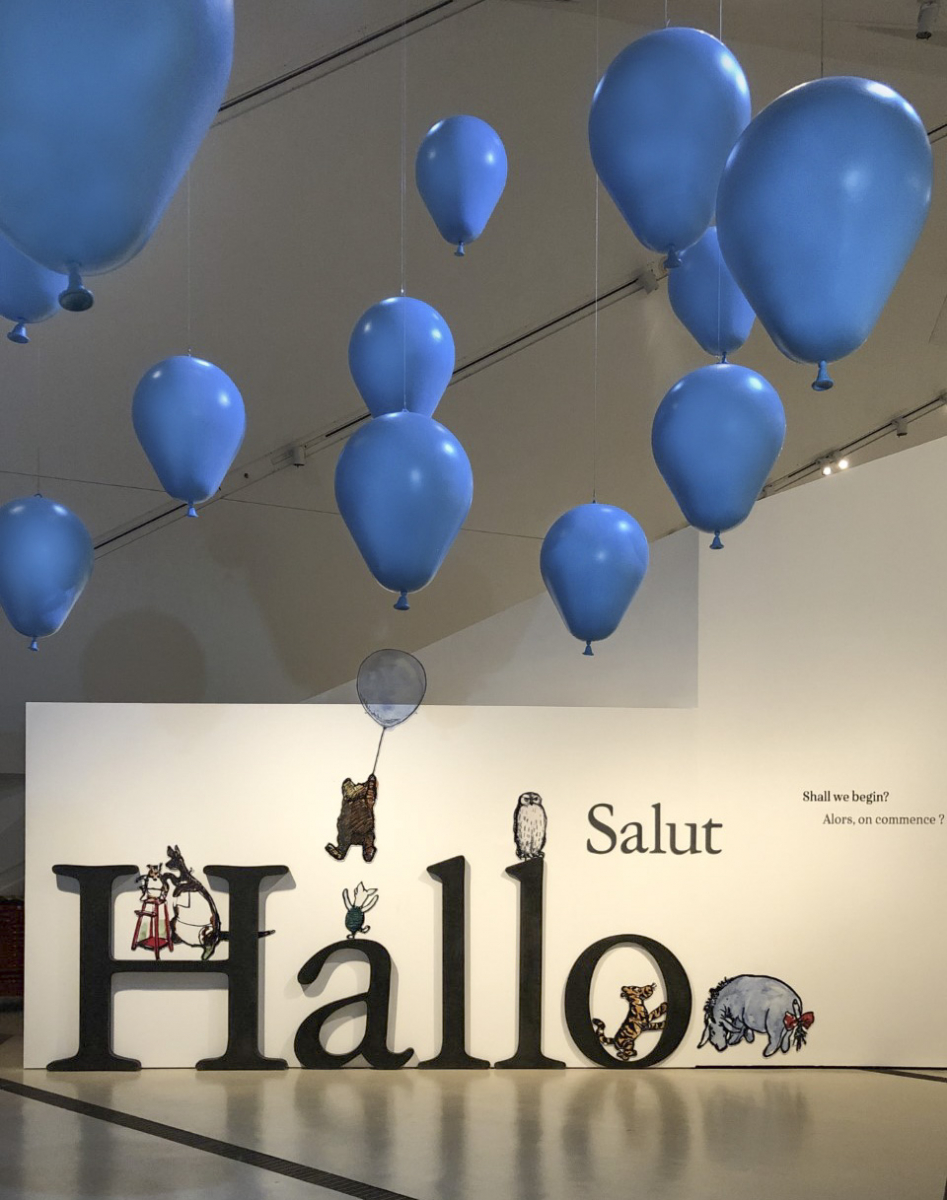 Hallo Salut in large black letters with small stuffed animal figures on and around the letters, and blue balloon-shaped sculptures floating above.
