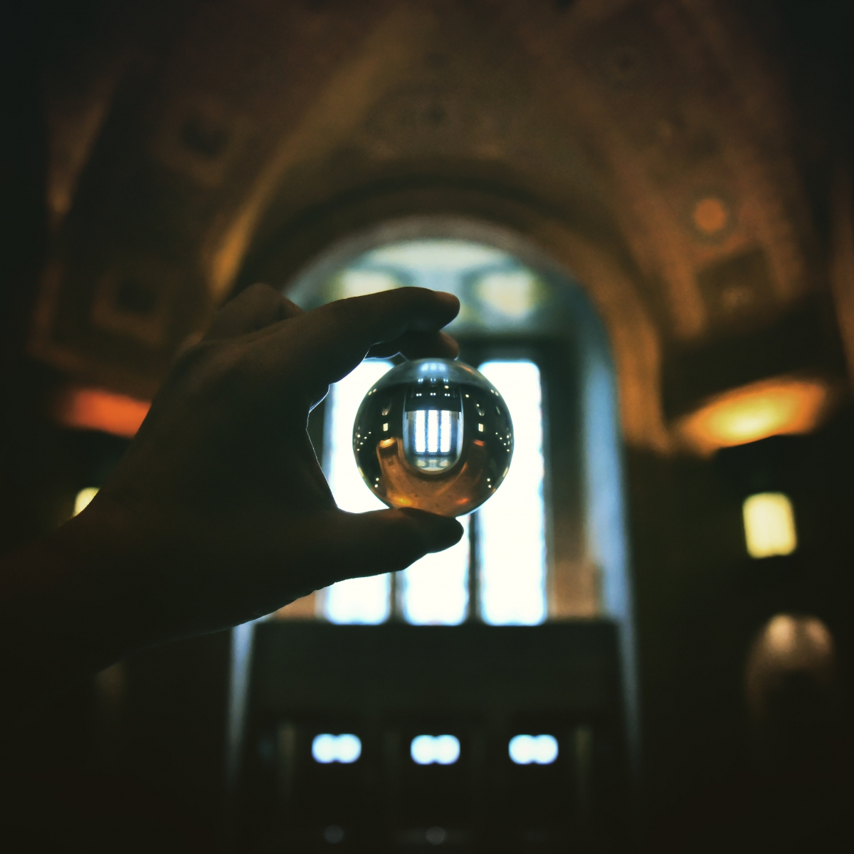 A hand holding a transparent sphere in front of an arched window with decorative wall sconces.