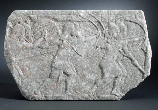 Stone carving depicting two figures, one riding an animal and the other holding a weapon, with incised details.