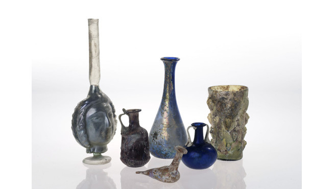 Five ancient glass vessels of different shapes and colors, including a tall blue vase, a small pitcher, a rounded bottle, a ribbed cup, and a small bird-shaped vessel.