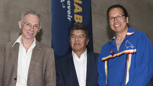 Three men standing side by side against a blue background, one wearing a brown jacket, one in a dark suit, and one in a blue outfit with colorful trim.