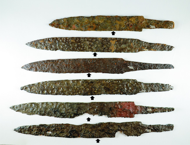 Six rusted iron spearheads with central holes, arranged vertically on a white background.