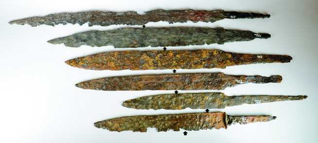 Seven rusted ancient iron spearheads of varying lengths arranged horizontally on a white background.