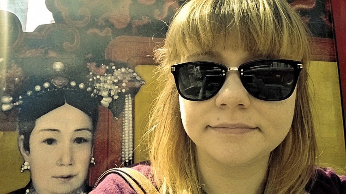 Woman with light brown hair wearing black sunglasses in front of a historical portrait of a woman with traditional Asian attire and headwear.