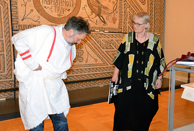 A man in a white tunic with red trim bends forward, while a woman in a black dress with colorful geometric patterns holds a stick and points.
