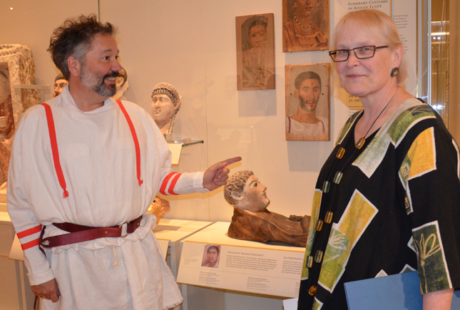 Two people stand inside a museum gallery with bust sculptures displayed on the wall and a pedestal.