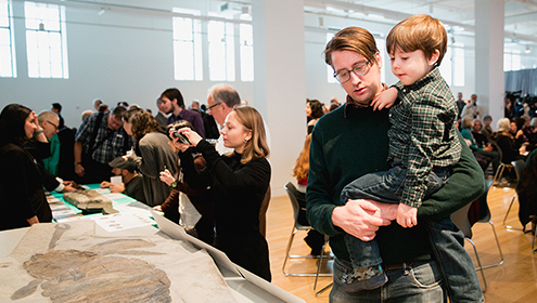 A woman examines an ancient sandy-colored artifact while a man carrying a young child looks on in a bright gallery space.