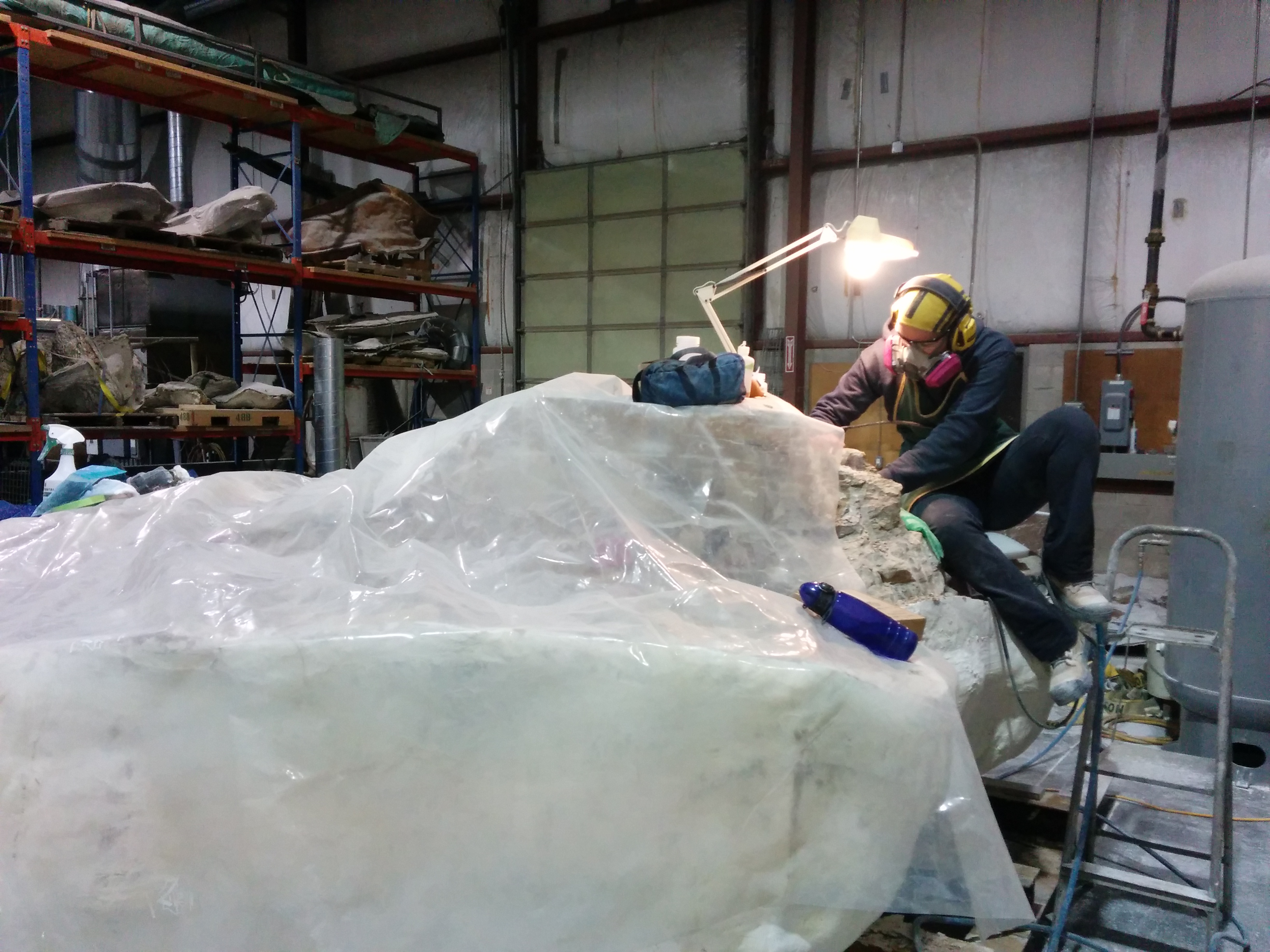 Two people wearing protective gear sculpting a large ice block inside an industrial workshop.