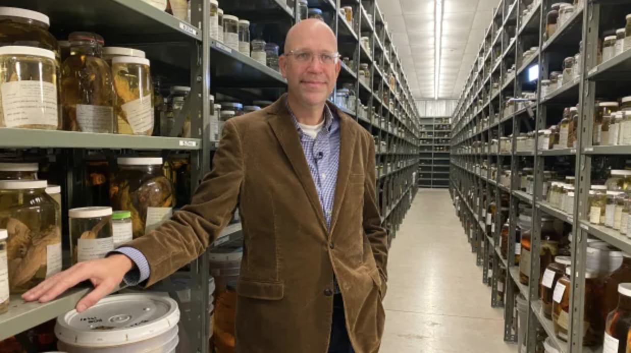 A man wearing glasses, a brown jacket, and a blue shirt stands in a narrow aisle lined with shelves stocked with jars and containers.