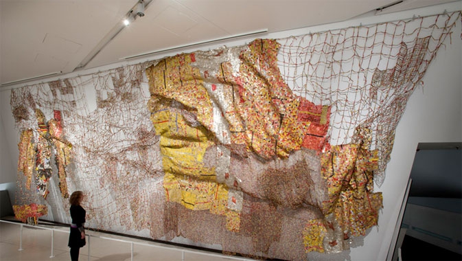 Large wall-mounted map artwork made from netting, layered fabrics, and various textures in brown, yellow, orange, and red tones.