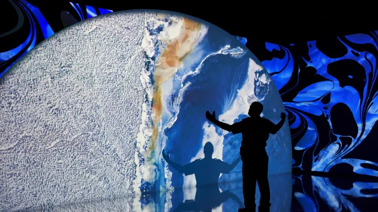 Projection of a planet with a silhouette of a man with his hands in the air looking at a large projection of a planet