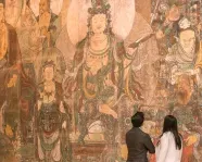 Two people looking at a large, detailed mural depicting multiple Asian-style figures wearing traditional robes.
